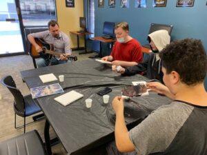 Students working at table while adult plays guitar in background