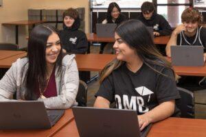 Students working in rows of desks with laptops. Front two women smiling at each other
