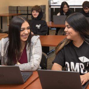 Students working in rows of desks with laptops. Front two women smiling at each other